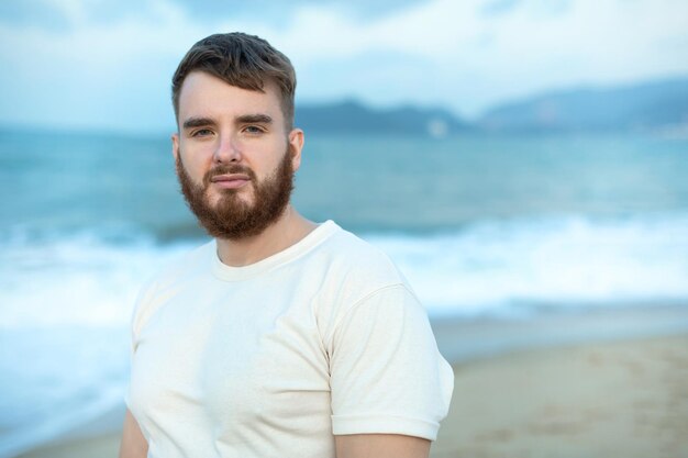 Ritratto di un giovane uomo felice e barbuto, un bel uomo sul mare o sull'oceano, che cammina sulla spiaggia e si diverte.