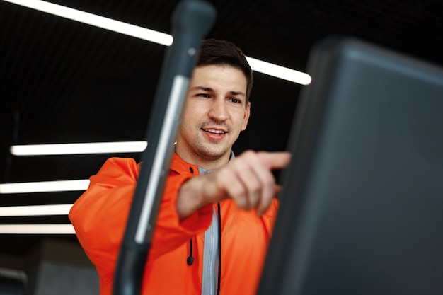 Ritratto di un giovane uomo in giacca a vento arancione allenamento su una macchina per il fitness in palestra