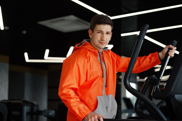 Ritratto di un giovane uomo in giacca a vento arancione allenamento su una macchina per il fitness in palestra