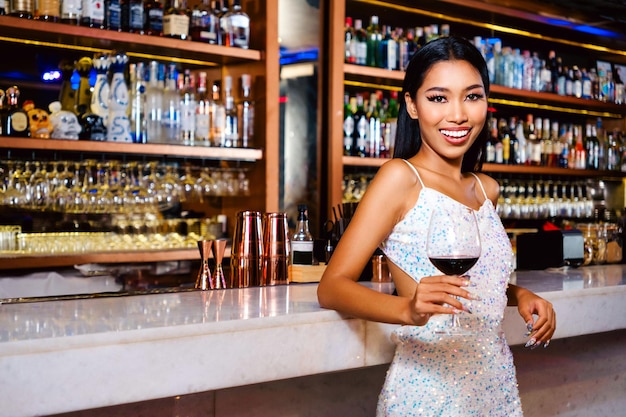 Ritratto di un glamour asiatico che indossa un vestito bianco luccicante sorridendo e bevendo un bicchiere di vino