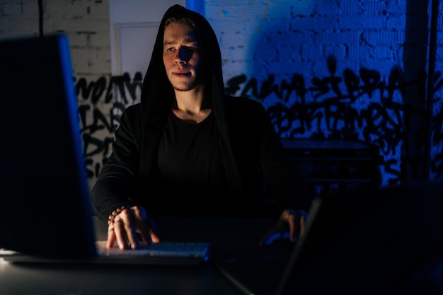Ritratto di un hacker nascosto che indossa una felpa con cappuccio impegnato nell'hacking nei sistemi di sicurezza seduto in una stanza buia nel seminterrato con luci al neon blu