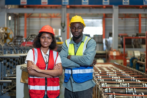 Ritratto di un ingegnere afroamericano femminile e maschile che indossa un giubbotto di sicurezza con casco sorridente con le braccia incrociate e guardando la fotocamera in piedi mentre lavora in fabbrica