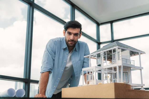 Ritratto di un ingegnere intelligente in abito casual che sorride alla telecamera mentre ispeziona il modello di casa Un architetto esperto che guarda la telecamera e si trova vicino al progetto di modello di casa modello architettonico Tracery