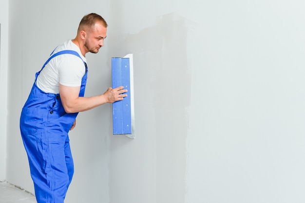 Ritratto di un lavoratore in tuta e con in mano una spatola sullo sfondo del muro intonacato. Lavori di riparazione e concetto di costruzione