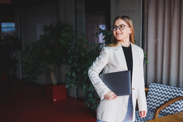 Ritratto di un manager di successo in ufficio che sorride durante la giornata lavorativa donna felice con il laptop che si gode il tempo in un ufficio moderno Donna bionda con una giacca beige e occhiali