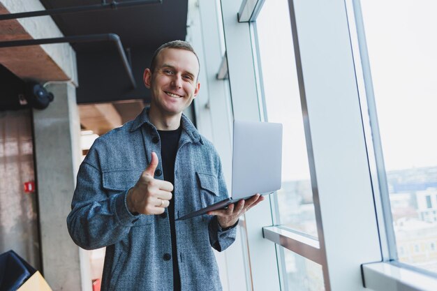 Ritratto di un manager maschio intelligente intelligente che tiene in mano un laptop per la ricerca in ufficio un uomo d'affari in abiti eleganti che gioca sul touchpad In piedi in ufficio vicino a una grande finestra