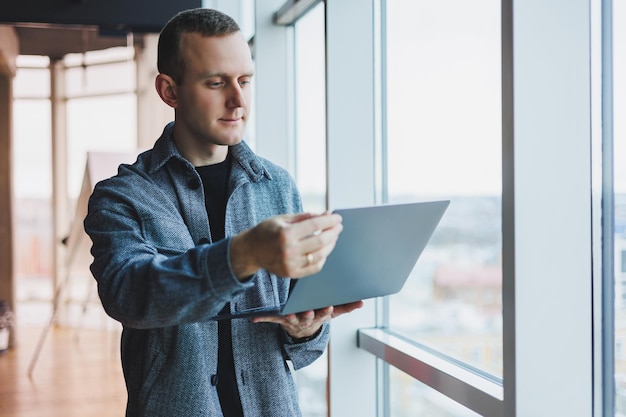 Ritratto di un manager maschio intelligente intelligente che tiene in mano un laptop per la ricerca in ufficio un uomo d'affari in abiti eleganti che gioca sul touchpad In piedi in ufficio vicino a una grande finestra