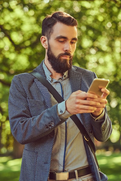 Ritratto di un maschio barbuto con un taglio di capelli elegante in abiti casual, scrive un messaggio al telefono, si trova in un parco cittadino.