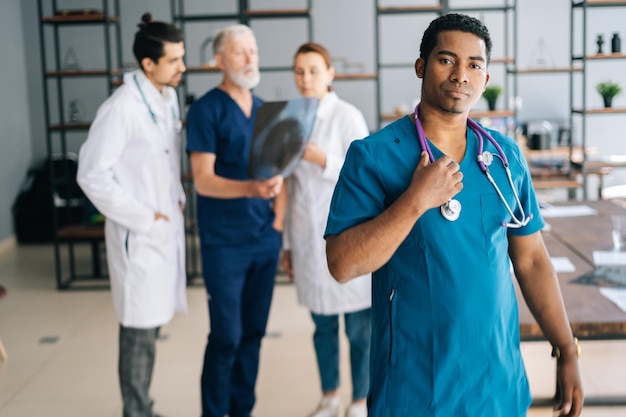 Ritratto di un medico maschio afroamericano serio in uniforme blu in piedi nell'ufficio della riunione medica che guarda l'obbiettivo