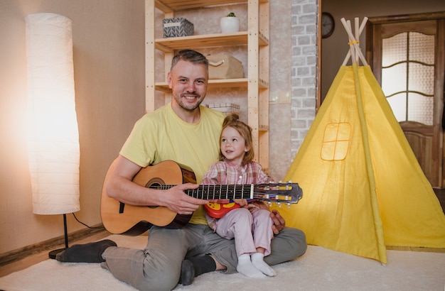 Ritratto di un padre felice con una chitarra in mano e una bambina seduta sulle ginocchia di suo padre. giochi familiari comuni.