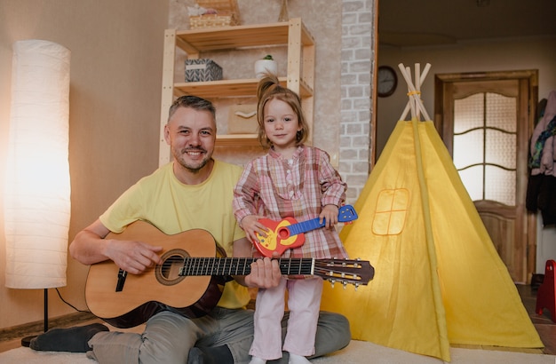 Ritratto di un padre felice con una chitarra in mano e una bambina seduta sulle ginocchia di suo padre. giochi familiari comuni.