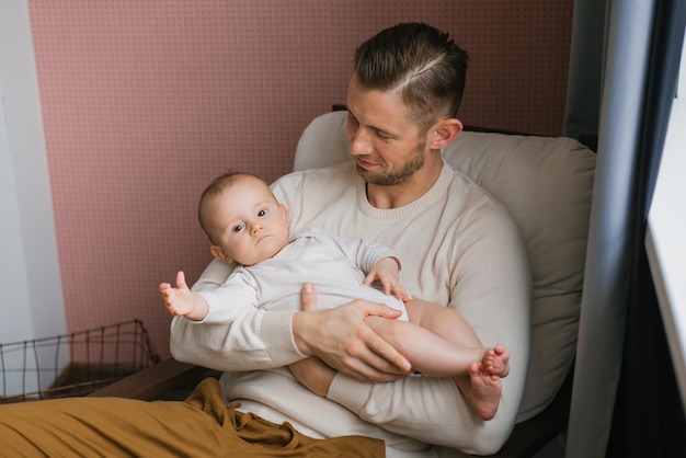 Ritratto di un papà caucasico premuroso che tiene un bambino carino tra le braccia seduto su una sedia comoda