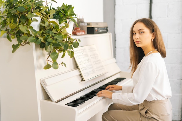 Ritratto di un pianista piuttosto femminile serio che suona su un pianoforte classico bianco in una stanza luminosa che guarda l'obbiettivo