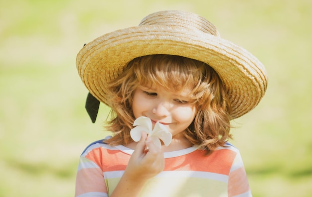 Ritratto di un piccolo ragazzo biondo con cappello di paglia in primo piano Viso carino per bambini con fiore di plumeria Bambino emotivo positivo nel parco naturale estivo Vacanze estive