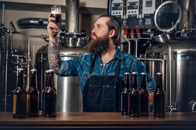 Ritratto di un produttore maschio hipster tatuato e barbuto che assaggia birra nel microbirrificio.