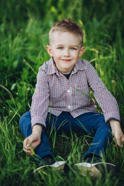 Ritratto di un ragazzino biondo alla moda che indossa una camicia a scacchi e jeans seduto nel parco in estate tra l'erba Bel ragazzo che gioca e sorride alla telecamera Look e moda per bambini