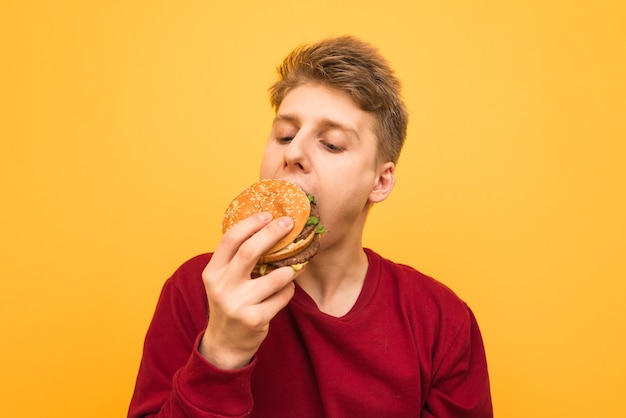 Ritratto di un ragazzo affamato in abbigliamento casual morde un appetitoso hamburger su un giallo