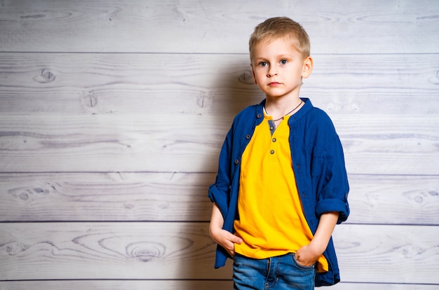 Ritratto di un ragazzo bellissimo bambino in maglietta gialla e giacca di jeans, camicia. Ragazzo che sta su un fondo di legno bianco.