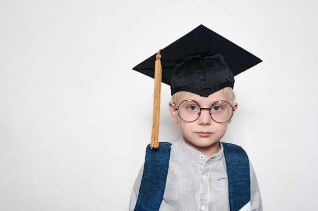 Ritratto di un ragazzo biondo carino in grandi occhiali, cappello accademico e una borsa di scuola.