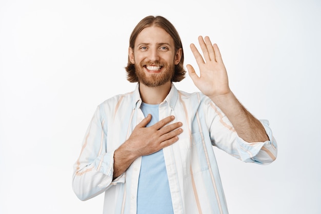 Ritratto di un ragazzo biondo sorridente amichevole che alza la mano, sembra felice, fa una promessa, si presenta, indossa una camicia, in piedi su bianco.