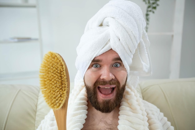 Ritratto di un ragazzo divertente che indossa un asciugamano turbante che tiene in mano la spazzola per massaggi mentre si siede sulla pelle maschile del divano