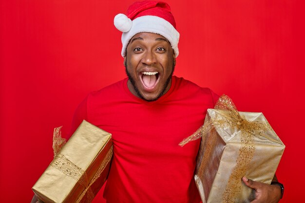 Ritratto di un ragazzo nero con un sorriso a trentadue denti con un cappello da Babbo Natale che tiene i regali di Natale sotto le ascelle