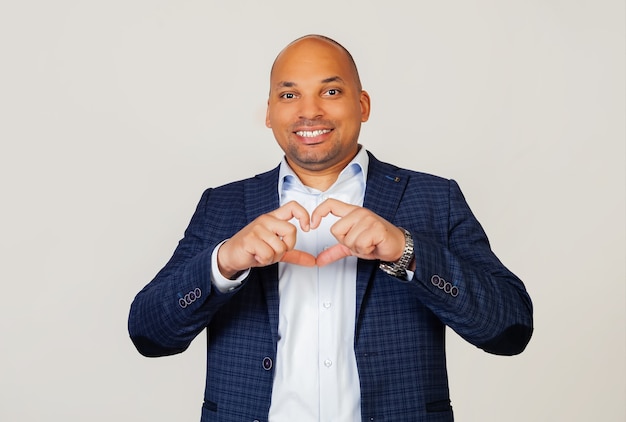 Ritratto di un ragazzo romantico giovane uomo d'affari afroamericano sorridente in amore che mostra il simbolo del cuore e la forma con le mani. Concetto romantico. In piedi su un muro grigio.