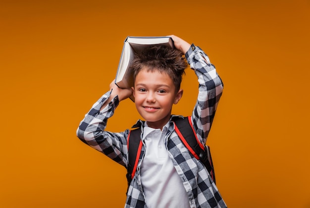 Ritratto di un ragazzo scolaro che tiene un libro sopra la testa con uno zaino a borsa isolato su uno sfondo giallo torna a scuola