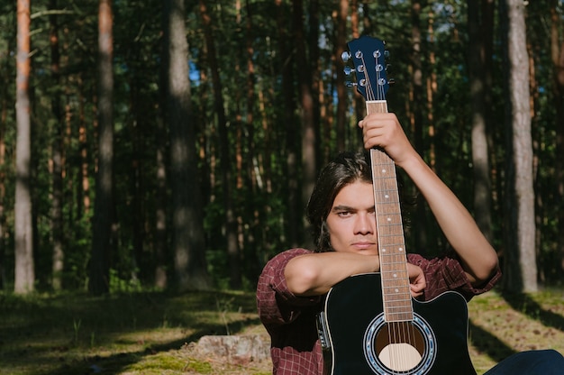 Ritratto di un ragazzo seduto nel bosco con una chitarra. Sullo sfondo del campo forestale.