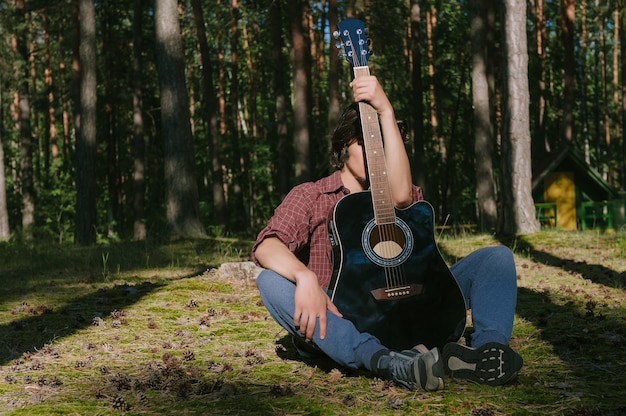 Ritratto di un ragazzo timido seduto nella foresta, coprendosi il viso con una chitarra. Sullo sfondo del campo forestale.