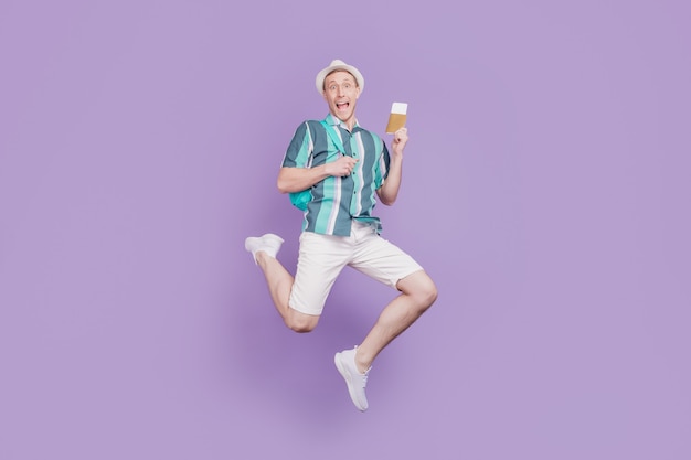 Ritratto di un ragazzo turistico positivo funky stupito che salta e tiene il passaporto dei biglietti su sfondo viola