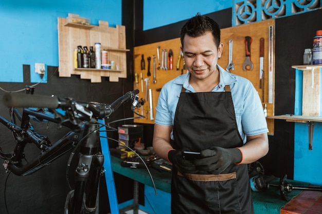 Ritratto di un riparatore in un grembiule che utilizza un cellulare mentre ripara una bicicletta