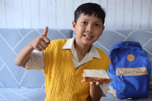 Ritratto di un simpatico ragazzino asiatico che mangia la colazione prima di andare a scuola e mostra il pollice in su