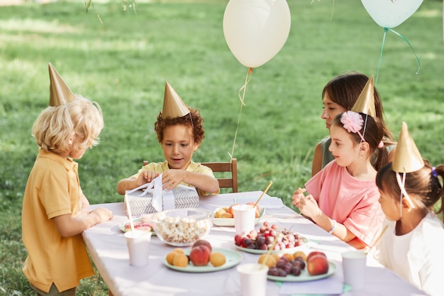 Ritratto di un simpatico ragazzo riccio che apre i regali durante la festa di compleanno all'aperto nello spazio estivo della copia