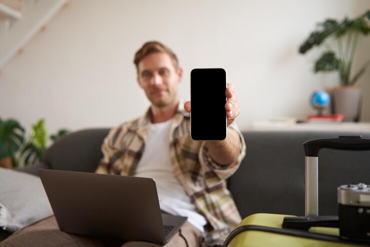 Ritratto di un turista che mostra lo schermo del suo cellulare e raccomanda un'app di viaggio seduto con una valigia