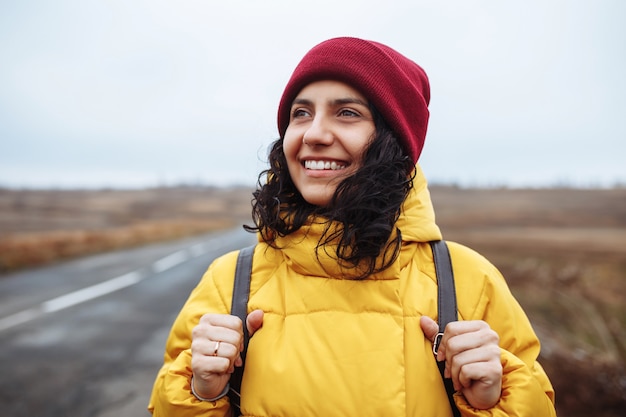 Ritratto di un turista femminile con uno zaino che indossa giacca gialla e cappello rosso si trova sulla strada.