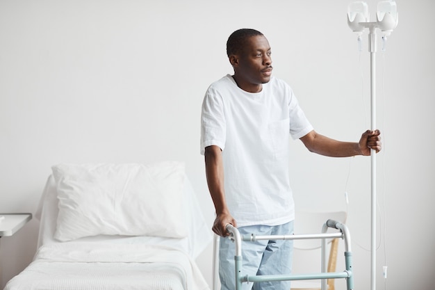 Ritratto di un uomo afro-americano in ospedale che tiene il supporto per flebo IV e distoglie lo sguardo pensieroso, copia spazio