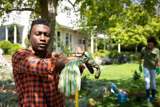 Ritratto di un uomo afroamericano in giardino, appoggiato a un palo con sua moglie sullo sfondo. Famiglia che si gode il tempo a casa, concetto di stile di vita