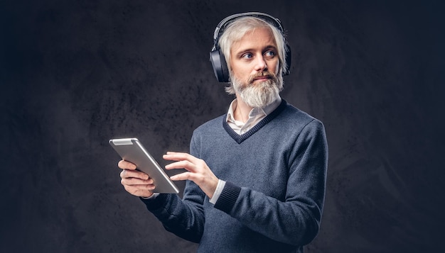 Ritratto di un uomo anziano bello che utilizza un tablet con le cuffie in uno studio su uno sfondo scuro.