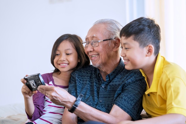 Ritratto di un uomo anziano felice e dei suoi nipoti che tengono insieme un telefono cellulare sul divano di casa