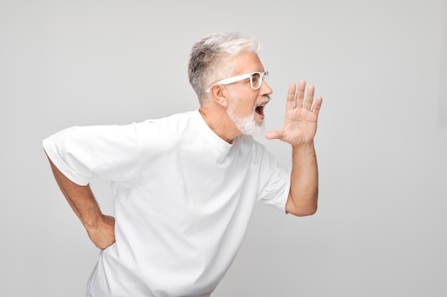 Ritratto di un uomo anziano in bianco che urla ad alta voce con le mani le palme delle notizie piegate come un megafono isolato su uno sfondo bianco