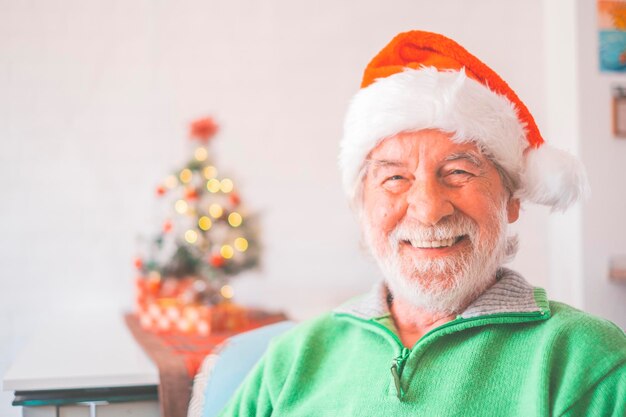 Ritratto di un uomo anziano in pensione sorridente con cappello da Babbo Natale e vestiti caldi che celebrano il natale a casa. Vecchio maschio santa sorridente mentre guarda la fotocamera. Uomo anziano che si gode le vacanze di Natale