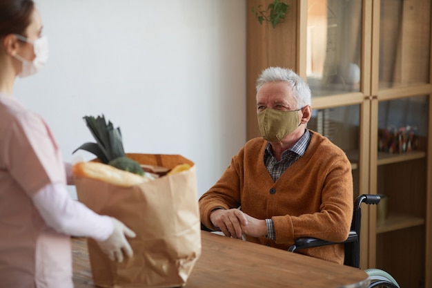 Ritratto di un uomo anziano sorridente in sedia a rotelle che parla con una donna che tiene in mano una borsa della spesa, assistenza e servizio di consegna cibo, spazio copia