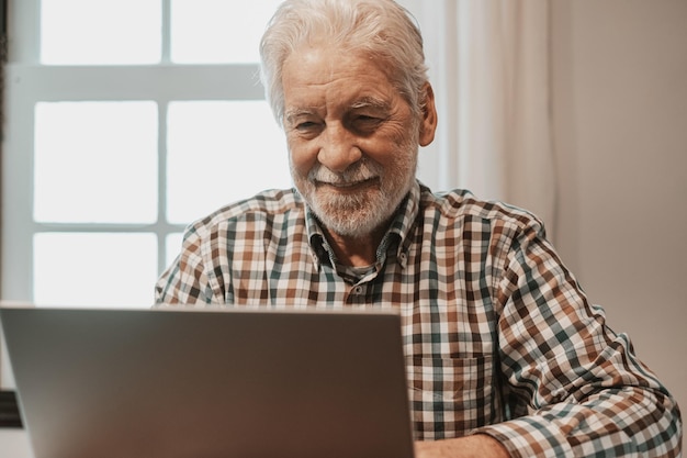 Ritratto di un uomo barbuto anziano sorridente seduto al tavolo con un computer portatile che naviga su siti online Uomo anziano in camicia a scacchi che utilizza le nuove tecnologie facendo acquisti o guardando video