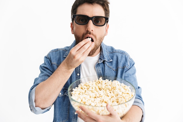 Ritratto di un uomo barbuto bello eccitato che indossa abiti casual in piedi isolato, guardando un film, mangiando popcorn