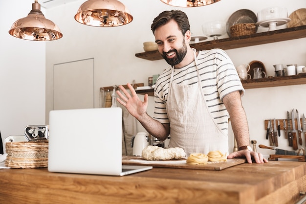 Ritratto di un uomo brunetta sorridente che indossa un grembiule usando il portatile mentre cucina e prepara la pasta fatta in casa in cucina a casa