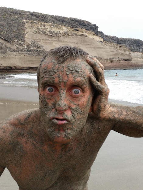 Ritratto di un uomo coperto di fango sulla spiaggia contro il cielo