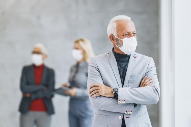 Ritratto di un uomo d'affari anziano pensieroso in piedi con le braccia incrociate in ufficio mentre indossa una maschera protettiva durante la pandemia del virus corona e guarda fuori dalla finestra.