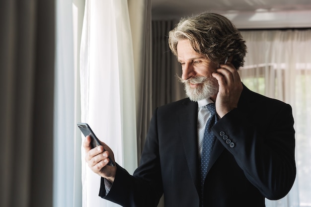 Ritratto di un uomo d'affari dai capelli grigi maturo barbuto serio in posa al chiuso a casa vicino alla finestra vestito in abiti formali utilizzando il telefono cellulare e gli auricolari.