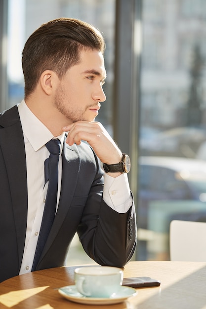 Ritratto di un uomo d&#39;affari serio bello che distoglie lo sguardo pensante sopra una tazza di caffè al caffè